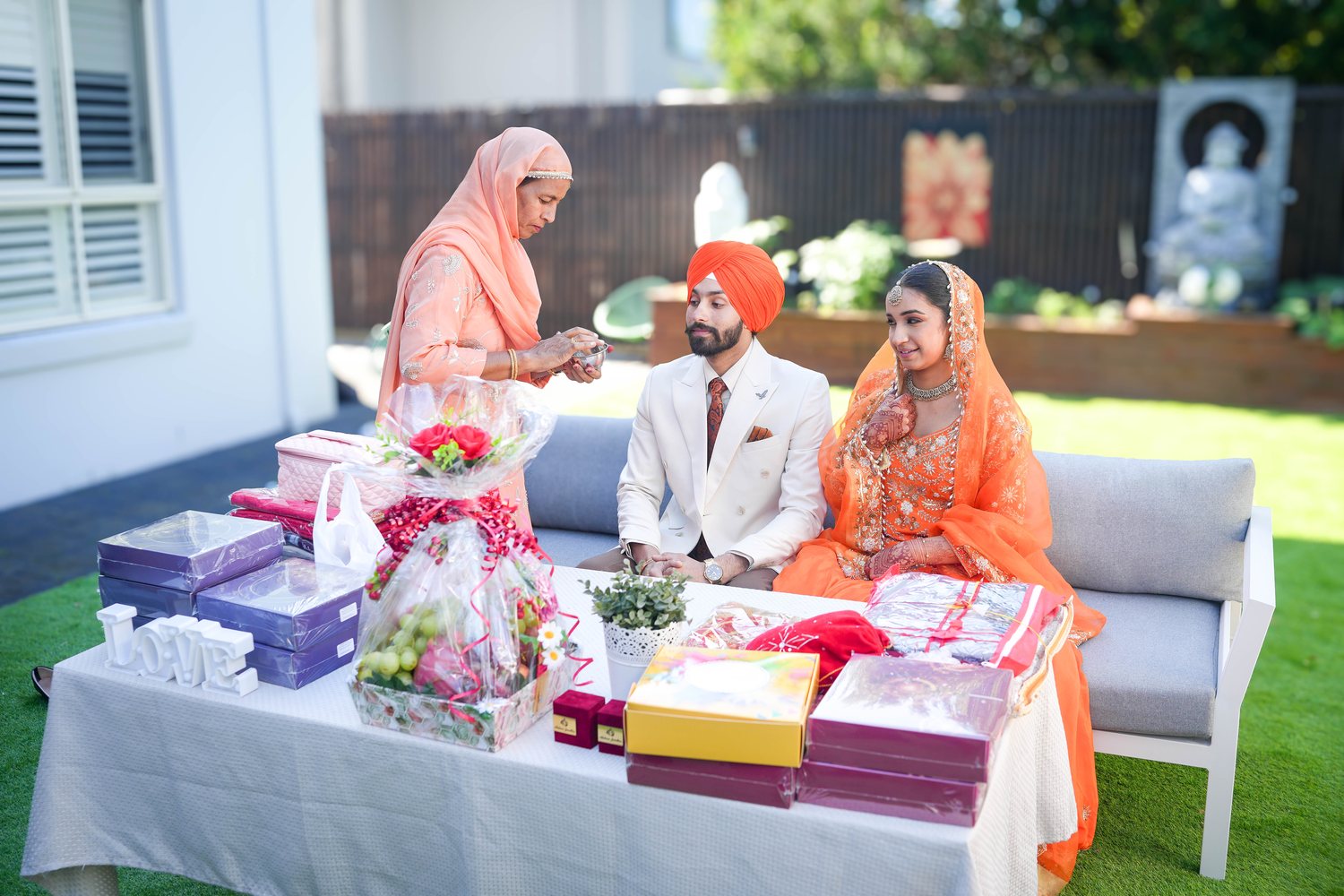 An elder performing a blessing ritual for a seated couple at a Sydney garden ceremony, gifts laid out on the table