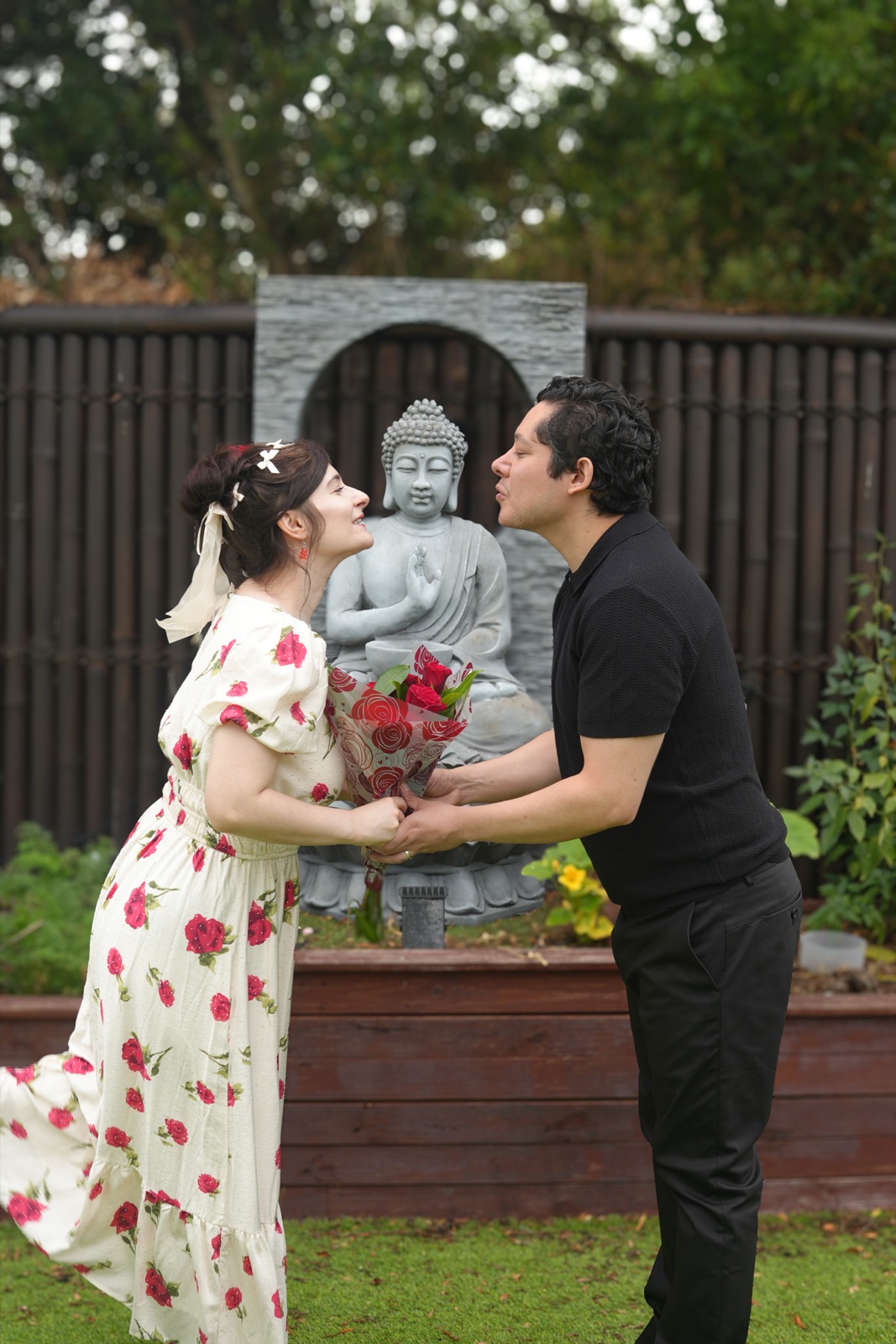 Manon and Karim leaning in to kiss, holding red roses, in front of a stone Buddha water feature in a Sydney garden