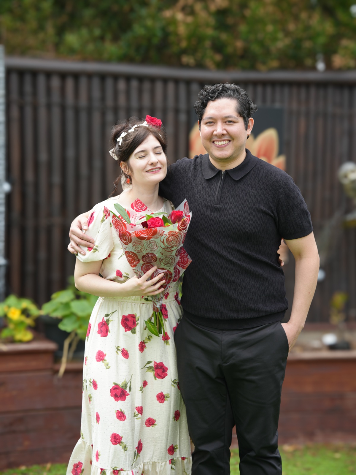 Karim with his arm around Manon, who leans into him holding a bouquet of red roses, in a Sydney garden