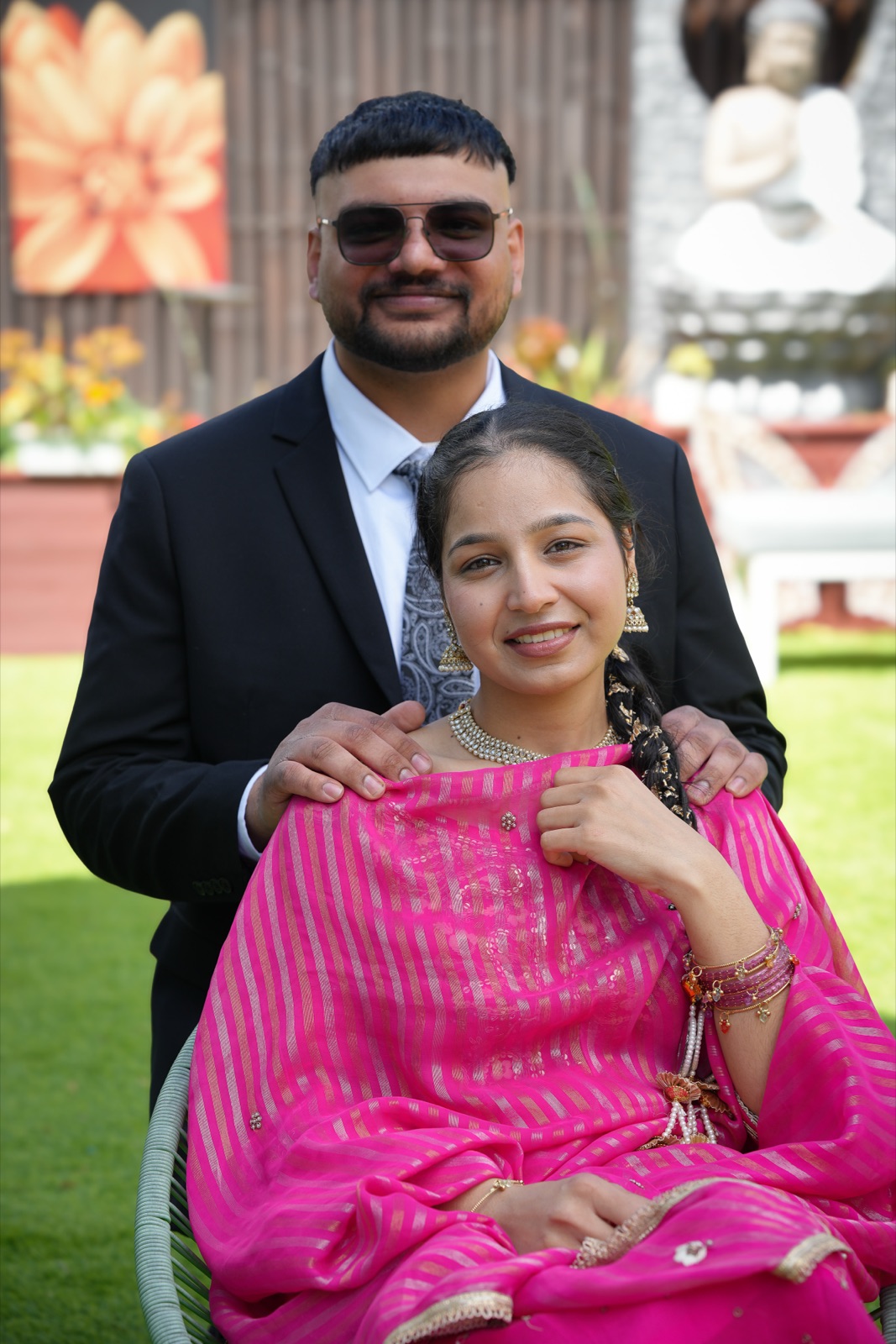 Gurpreet standing behind Harpreet, seated in a pink Punjabi suit and gold jewellery, at their Punjabi backyard wedding in Sydney