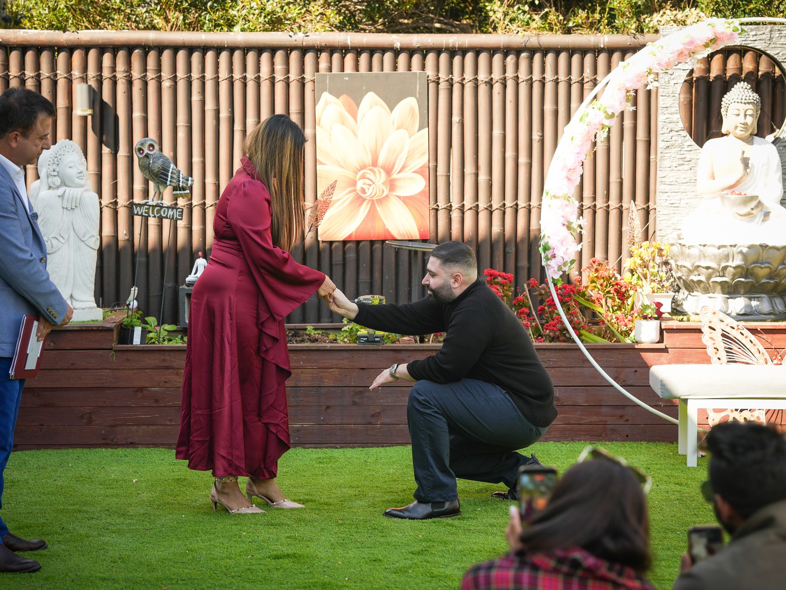 Groom kneeling for the ring exchange beneath a floral arch and Buddha statue at a Punjabi and Lebanese garden wedding in the Hills District, Sydney