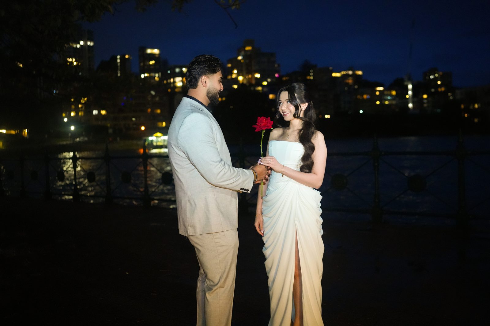 Couple exchanging a single red rose by Sydney Harbour at night, city lights behind them