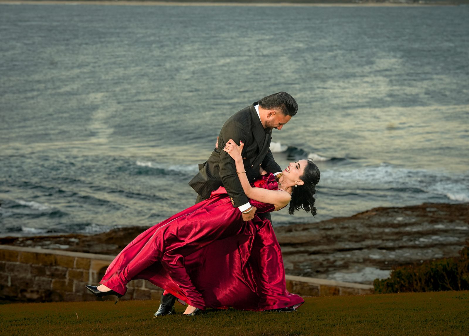Lovepreet dipping Gurpinder in a playful dance on the grass above the ocean at sunset after their Wollongong lighthouse wedding