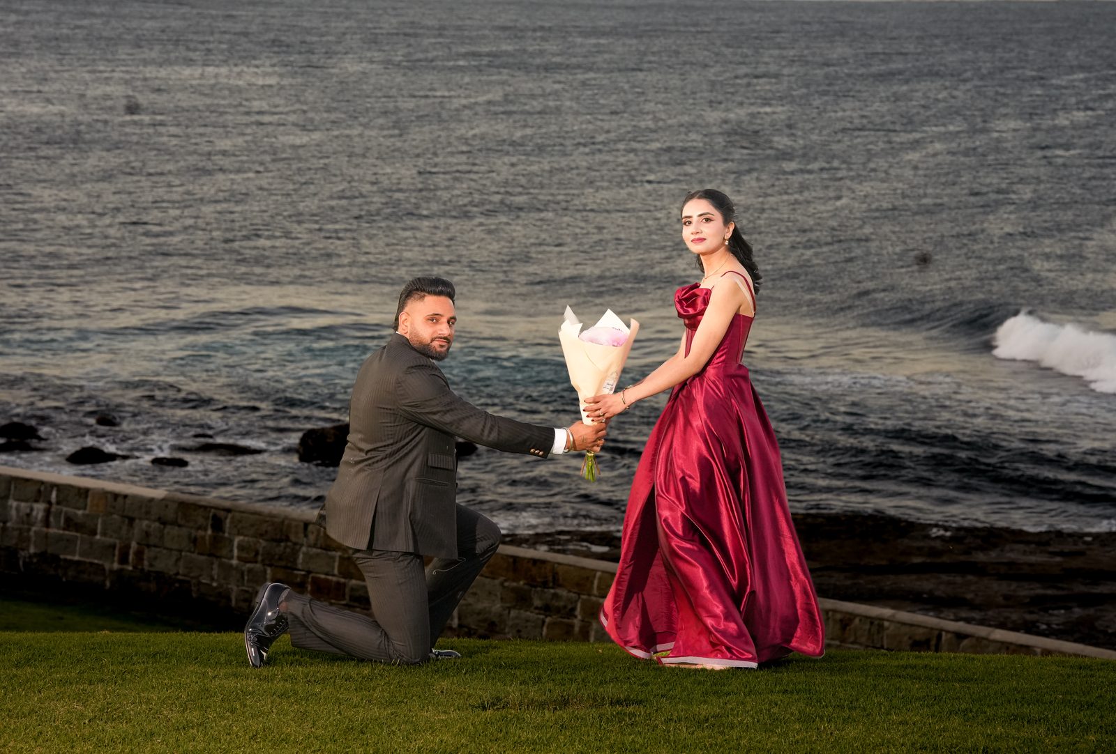 Lovepreet kneeling and offering a bouquet to Gurpinder beside the ocean at Wollongong lighthouse
