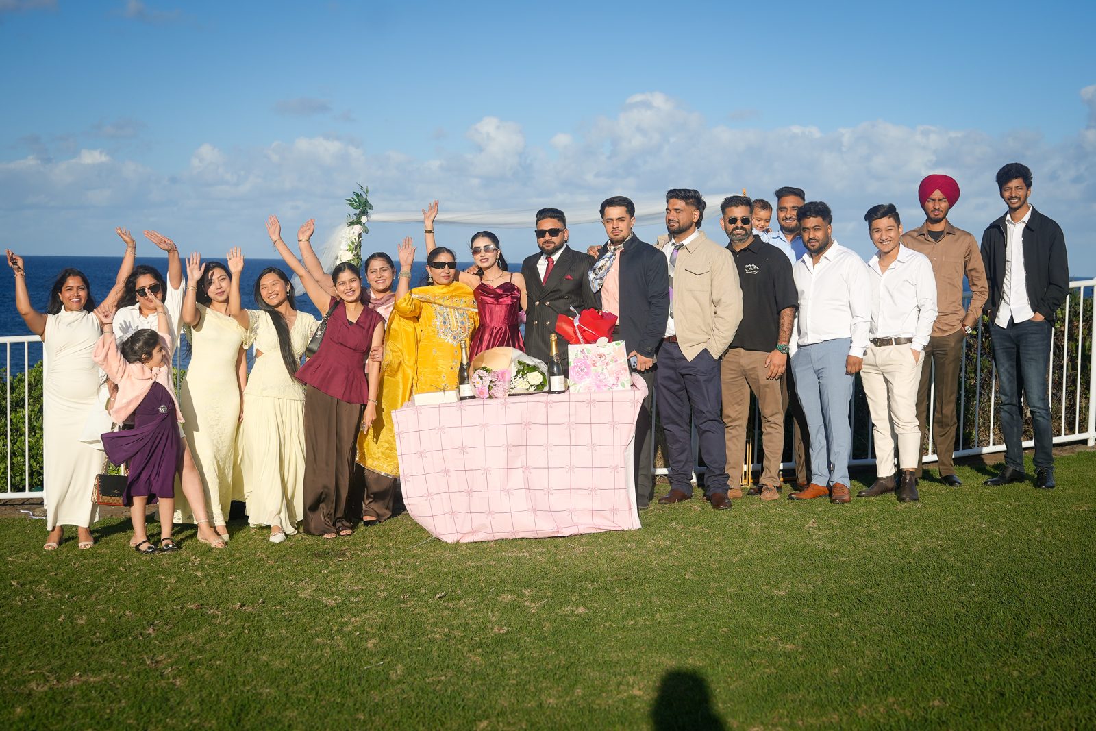 Lovepreet, Gurpinder and their guests cheering with raised arms beside a styled table on the grass at Wollongong lighthouse, with the ocean behind