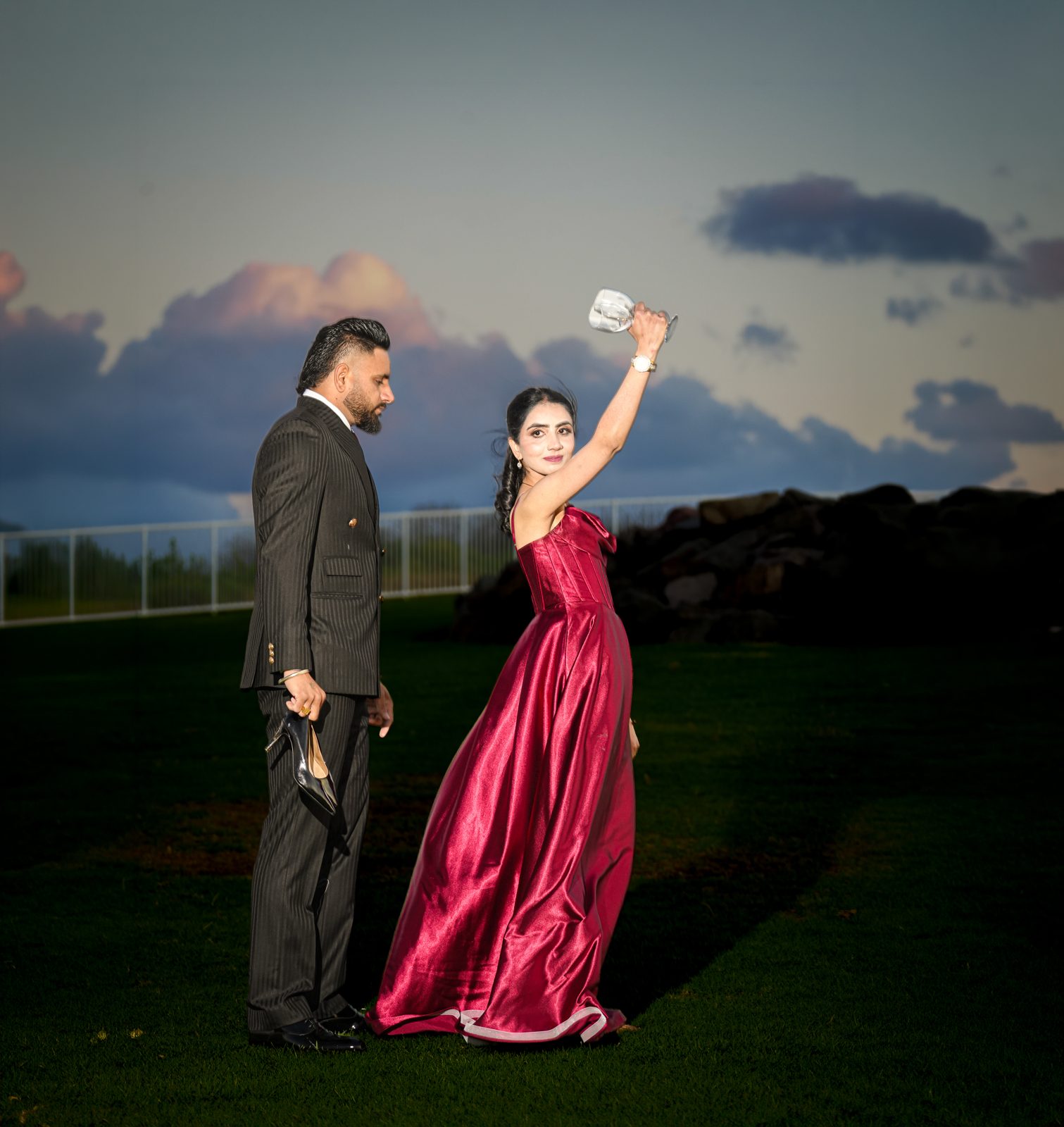 Gurpinder raising a glass and Lovepreet holding her shoes on the lawn at Wollongong lighthouse at dusk after their wedding